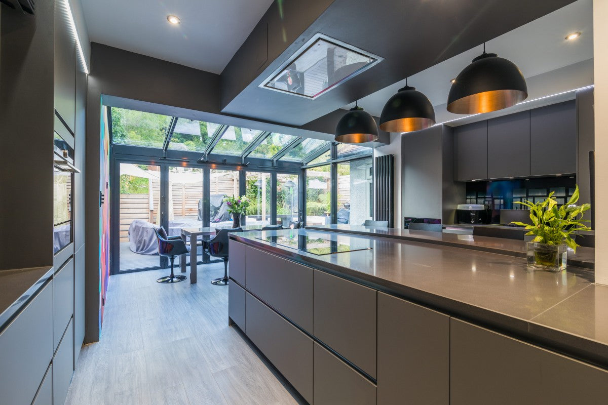 Long dark grey Silestone quartz kitchen island under copper pendant lights, opening onto a glass garden room